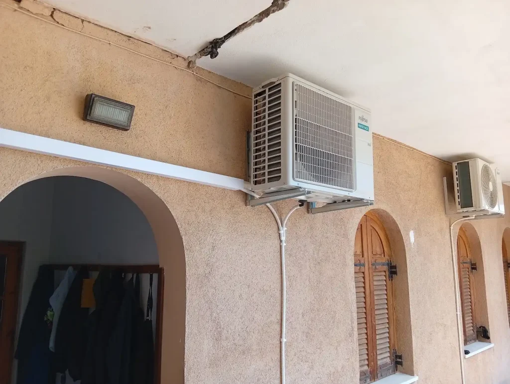 Εξωτερική μονάδα κλιματιστικού Fujitsu AIRSTAGE τοποθετημένη σε βάση στον εξωτερικό τοίχο κτιρίου, πάνω από αψιδωτά παράθυρα, με λευκό κανάλι προστασίας σωληνώσεων.