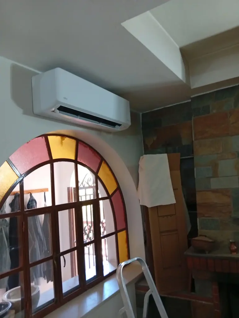 Λευκή εσωτερική μονάδα κλιματιστικού Fujitsu τοποθετημένη στον τοίχο πάνω από αψιδωτό παράθυρο με βιτρό, σε εσωτερικό χώρο με επένδυση πέτρας.