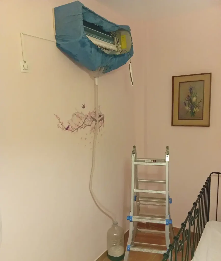 Εσωτερική μονάδα κλιματιστικού καλυμμένη με μπλε προστατευτικό κάλυμμα πλυσίματος και σωλήνα αποστράγγισης κατά τη διάρκεια συντήρησης, με μεταλλική σκάλα στο δωμάτιο.